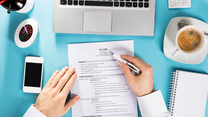 Resume being reviewed on blue desk with laptop and coffee. 