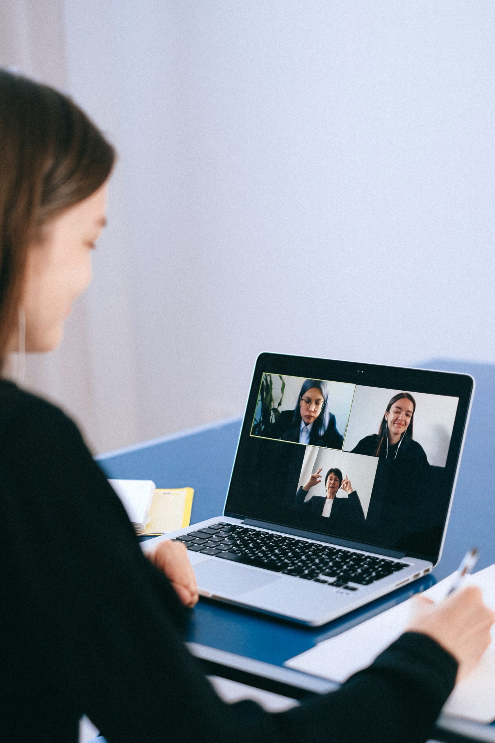 Open laptop displaying video call with three smiling professionals in front of professional taking notes. 